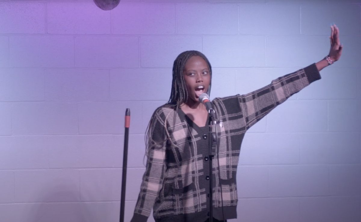 Performer in a plaid cardigan singing with one arm raised